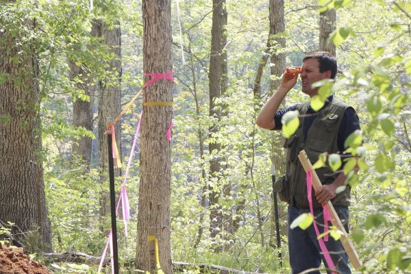 A soil engineer from Soils Inc. in the middle of a forest like area inspecting something by the hand lens